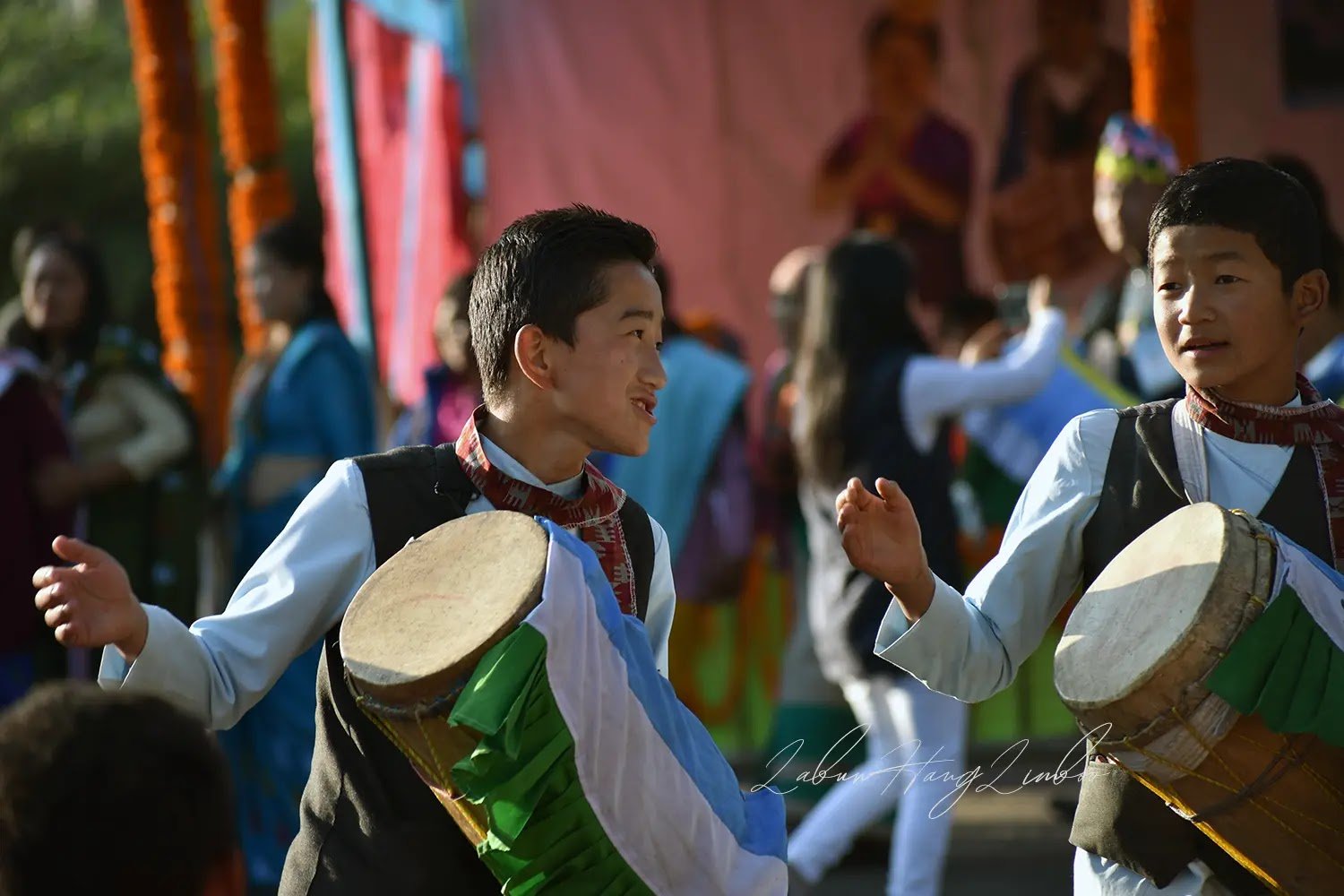 Two young Limbu men in traditional vests and scarves energetically perform the traditional Chyabrung dance (also known as Key Lang) at a cultural festival in Sikkim. They are smiling while playing the large, colorful Chyabrung drums, celebrating the rich indigenous culture of the Limbu community of Sikkim.